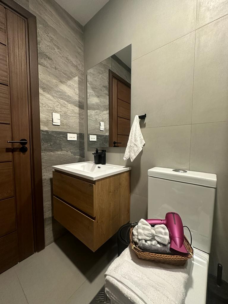 A modern bathroom features a sleek wooden vanity with an integrated sink and a large mirror. A white toilet is positioned beside the vanity. A towel hangs from a black rack, and essential toiletries are arranged in a woven basket on a soft towel.