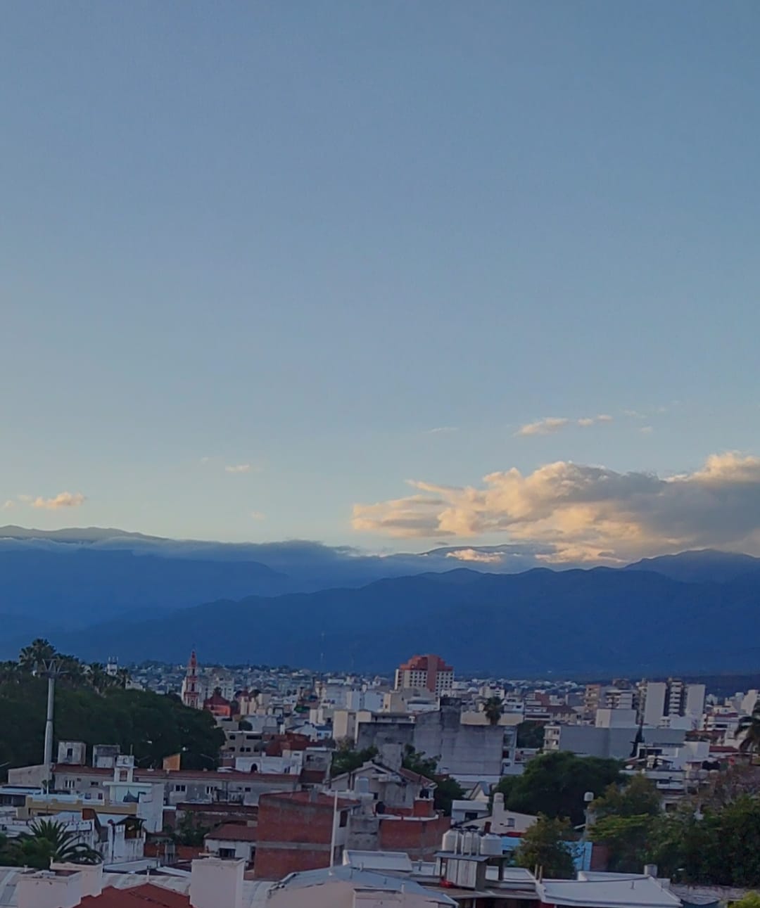 A panoramic view of the city is captured, featuring a mixture of buildings and rooftops under a clear sky. Majestic mountains rise in the background, with a few clouds drifting overhead, creating a tranquil landscape.