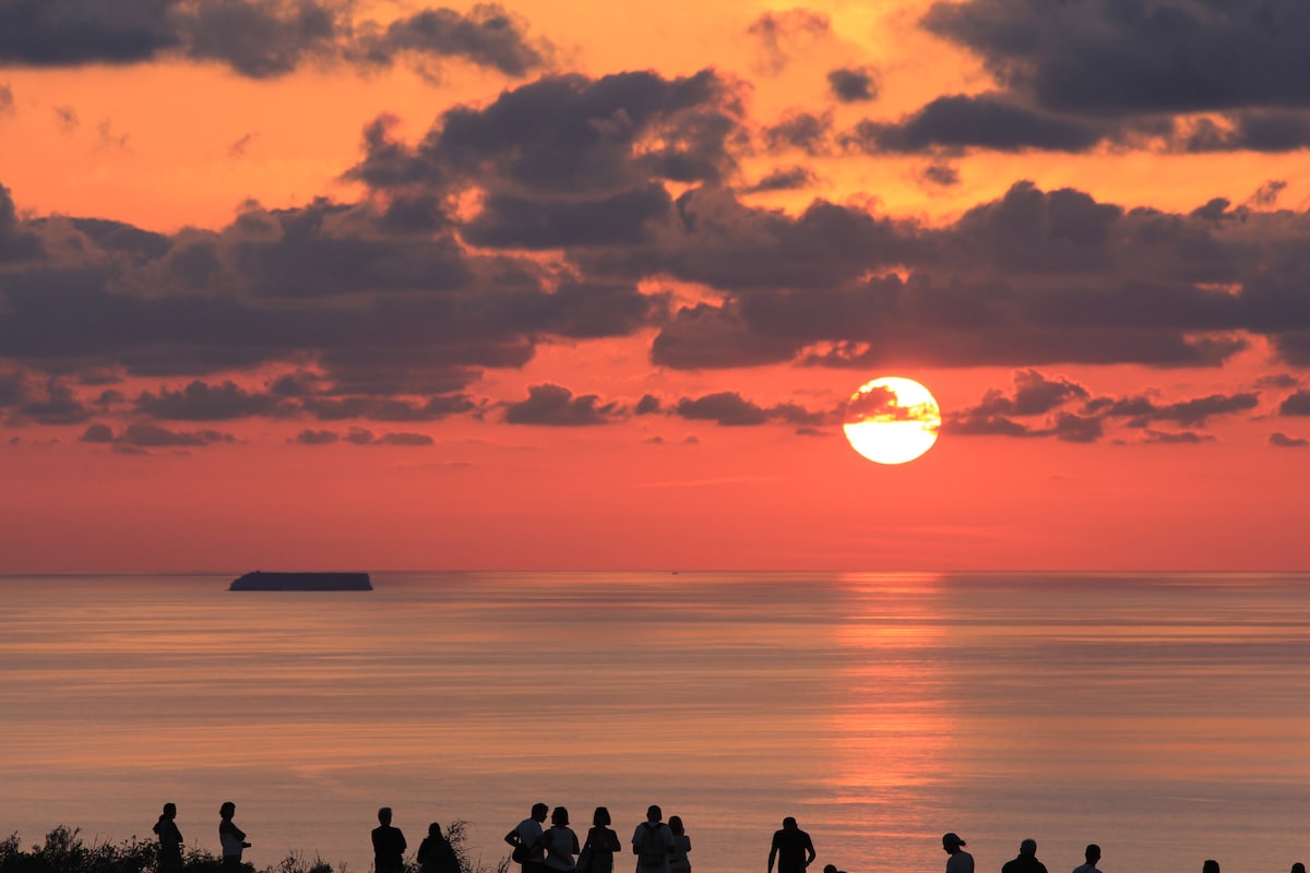 A sunset is captured over the calm sea, with vibrant hues of orange and pink reflecting off the water. Silhouetted figures can be seen gathering along the shoreline, watching as the sun sets behind distant clouds and an island on the horizon.