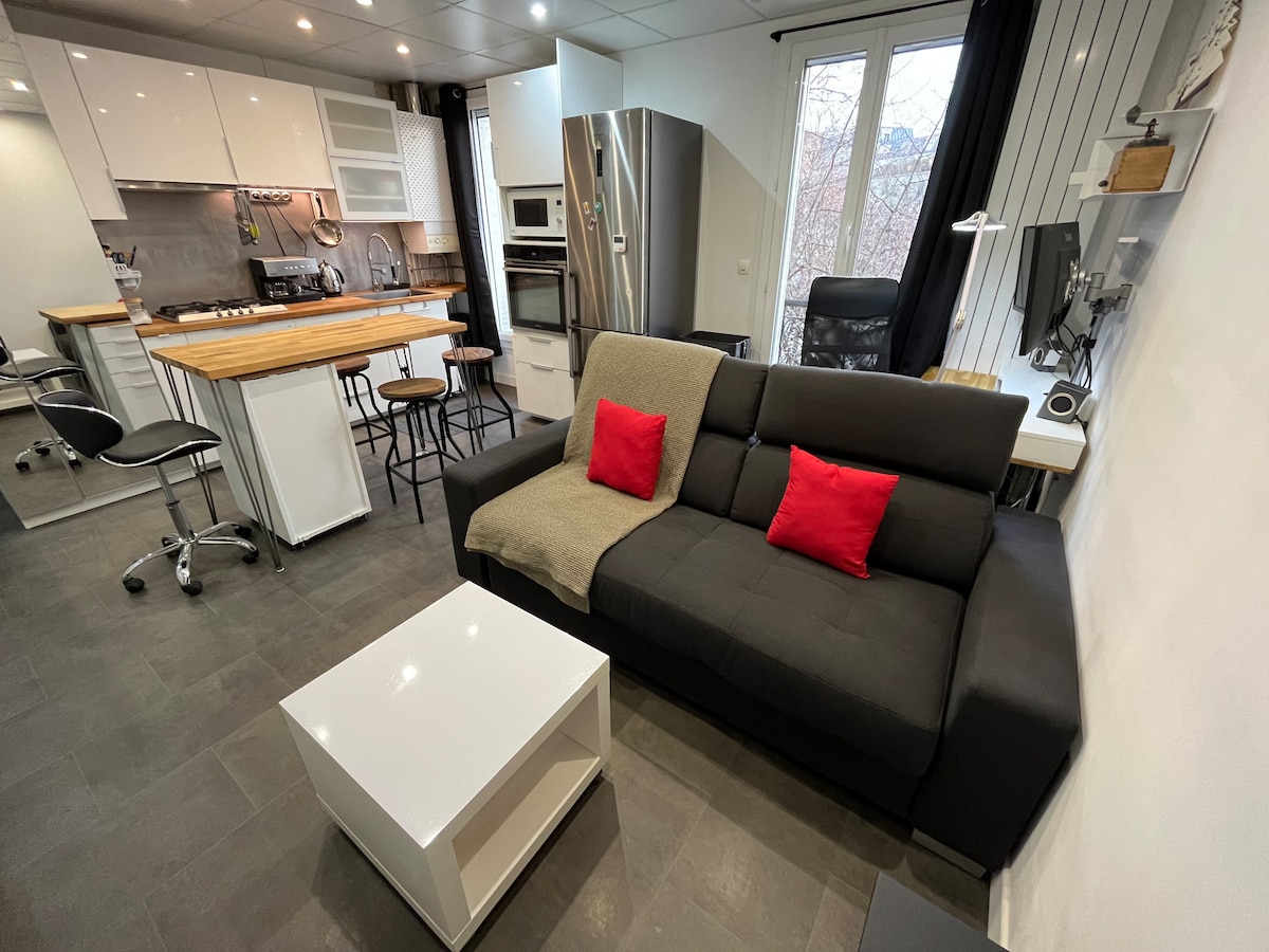 An open-plan kitchen and living area features a modern design with sleek cabinetry and appliances. A dark sofa with red accent pillows and a white coffee table complements the space. High-top seating circles a wooden kitchen island, providing ample room for socializing and dining.