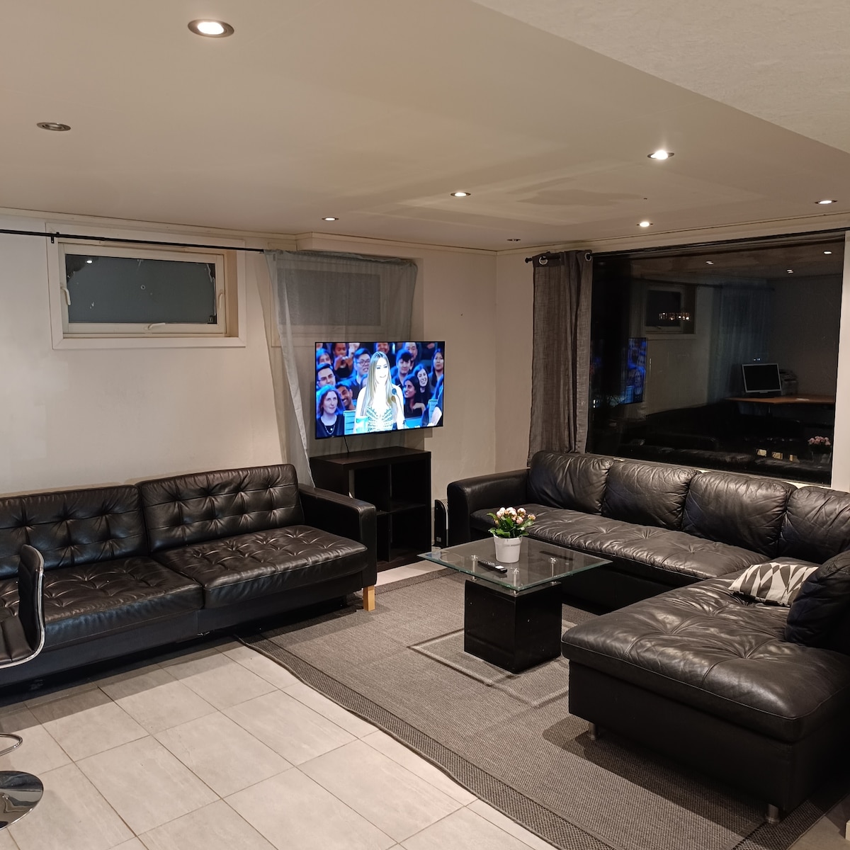 A modern living area features a black leather sectional sofa and a glass coffee table. A flat-screen television is positioned against the wall. Natural light enters through a window adorned with light curtains, while a minimalistic desk is visible in the background.