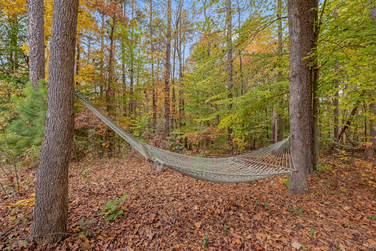 A hammock is gently suspended between two trees, surrounded by a forest of trees adorned with autumn leaves. The ground is covered with fallen leaves in various shades, creating a serene and inviting natural setting.