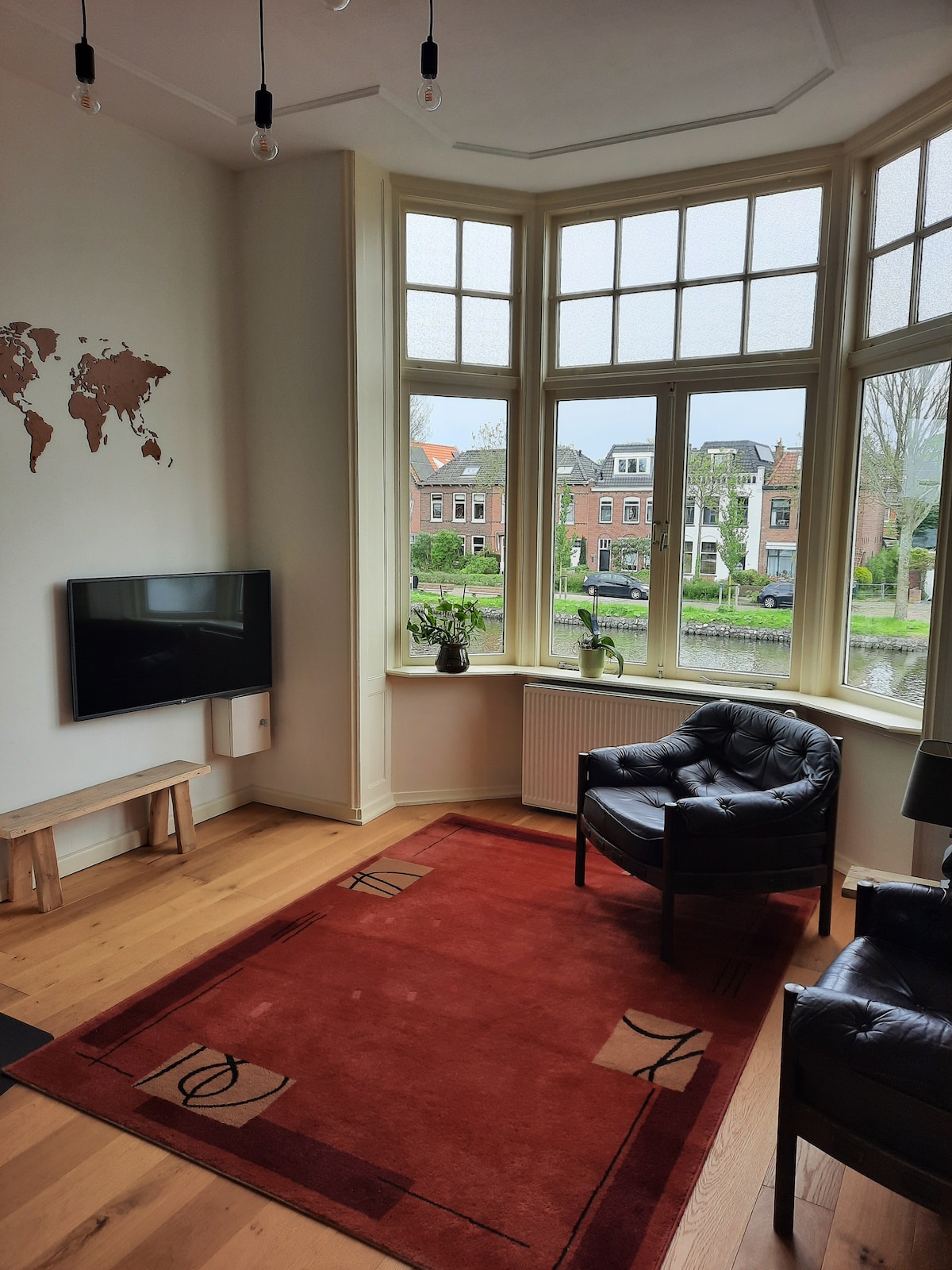 A corner seating area is highlighted by large windows allowing natural light to fill the space. A black armchair is positioned on a red area rug, with a wooden TV stand beneath a wall-mounted television. Potted plants adorn the windowsills, complementing the view of the street.