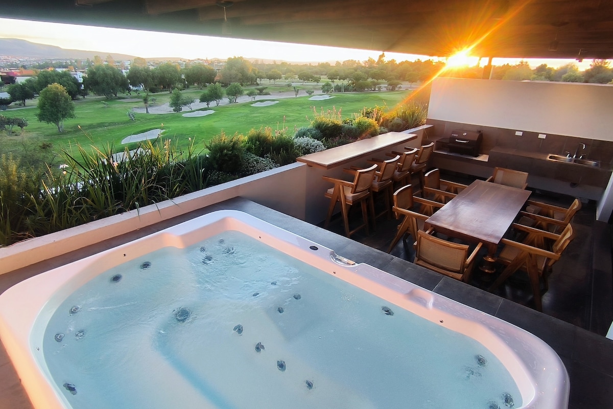 An outdoor terrace features a hot tub beside a long dining table made of wood. The area is surrounded by greenery and offers views of a well-maintained golf course, with the sun setting in the distance, casting a warm glow over the scene.