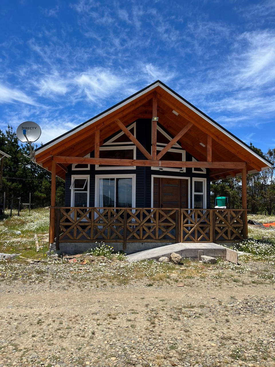 A charming cabin features a wooden porch with a decorative railing, set against a backdrop of blue skies and scattered clouds. The structure showcases exposed beams and large windows, allowing ample natural light. A satellite dish is mounted on the roof for connectivity.