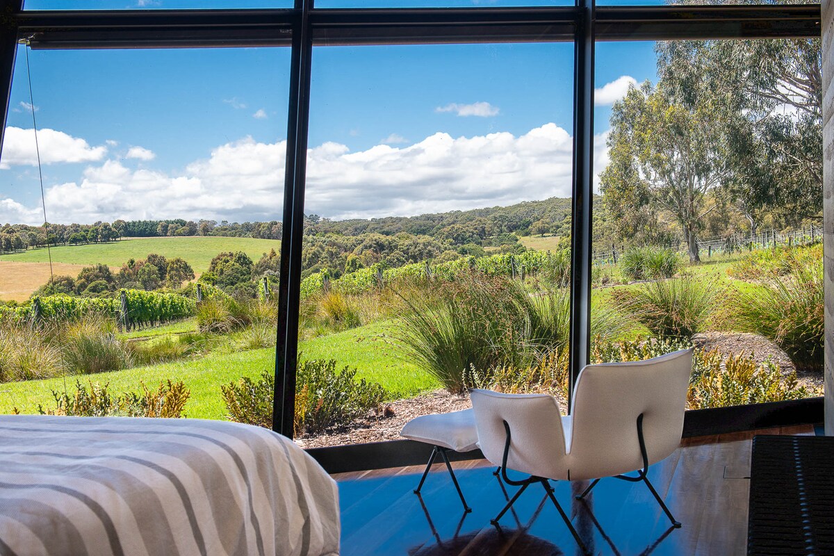A spacious bedroom is framed by large glass windows, providing a scenic view of the lush, green landscape and rolling hills. Two modern chairs are positioned near the windows, inviting relaxation while taking in the serene surroundings.
