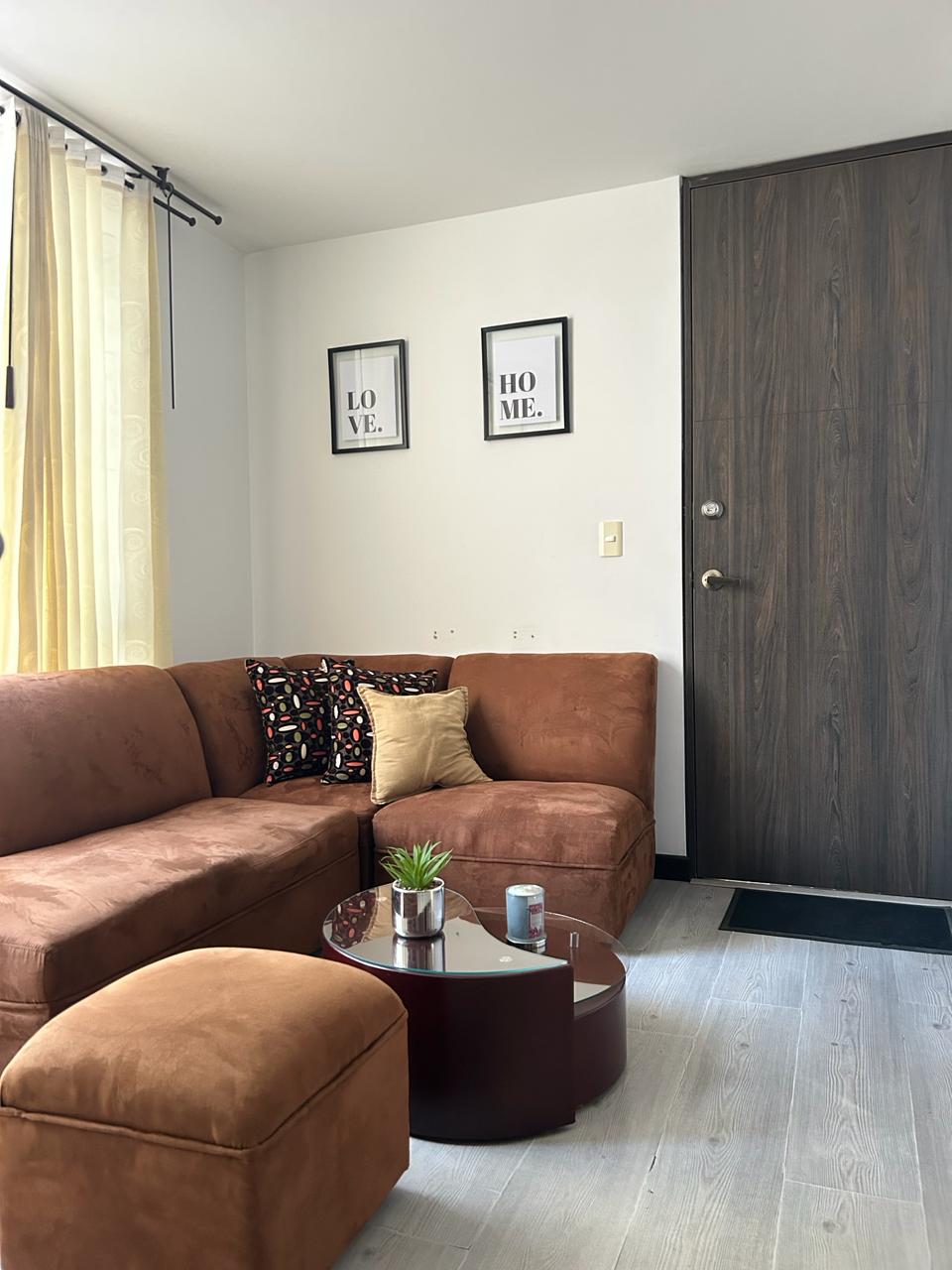 A living area features a brown upholstered sectional sofa with colorful accent pillows. A small round coffee table with a potted plant sits in the center. Two framed artworks adorn the wall above, while bright curtains allow natural light to fill the space.