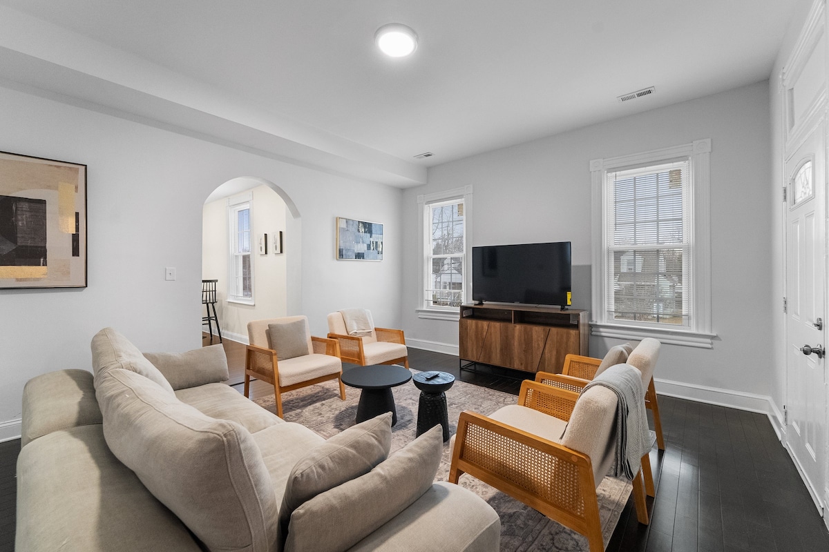 The spacious living area features a neutral color palette, complemented by large windows allowing natural light. A sectional sofa and four light wooden chairs surround a small table, while a modern television is positioned on a low wooden console. An archway leads to a hallway.