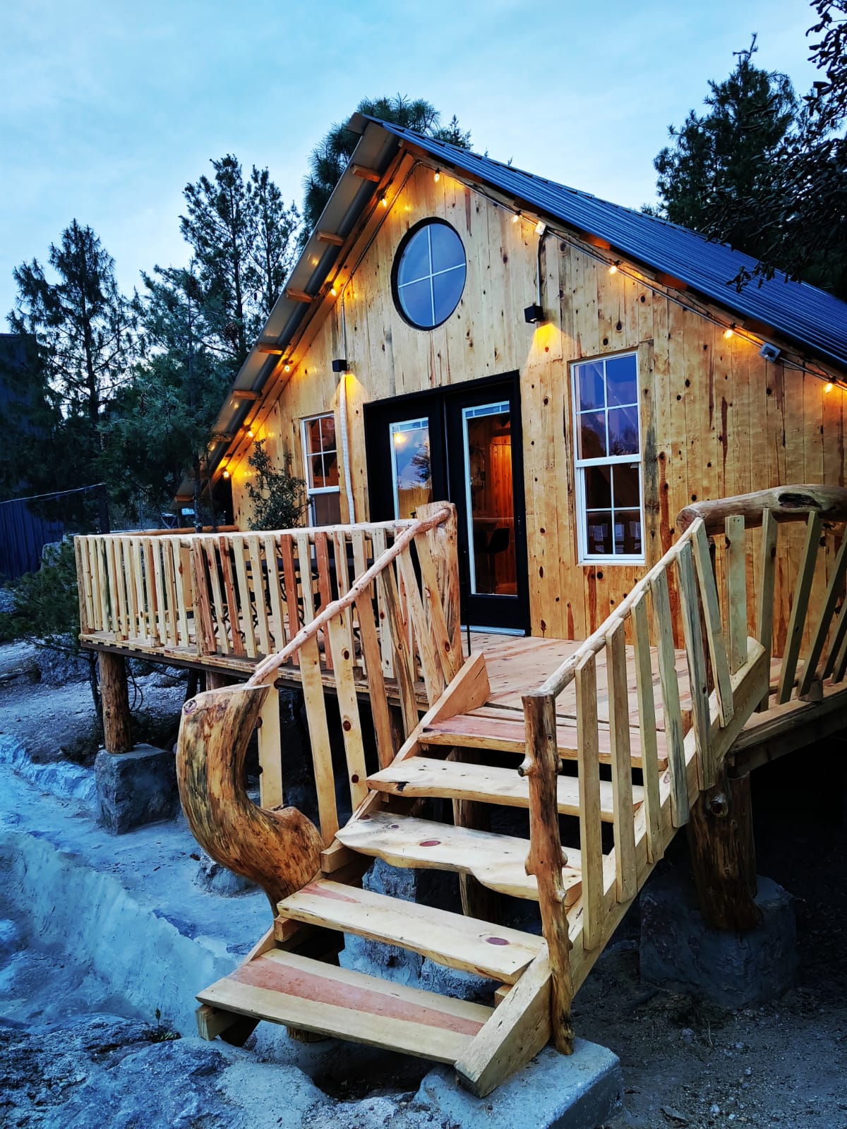A charming wooden cabin features a curved wooden staircase leading to the entrance, framed by decorative fairy lights. Large windows allow natural light to illuminate the interior, enhancing the cozy atmosphere. Surrounding trees provide a natural backdrop, contributing to a serene setting.