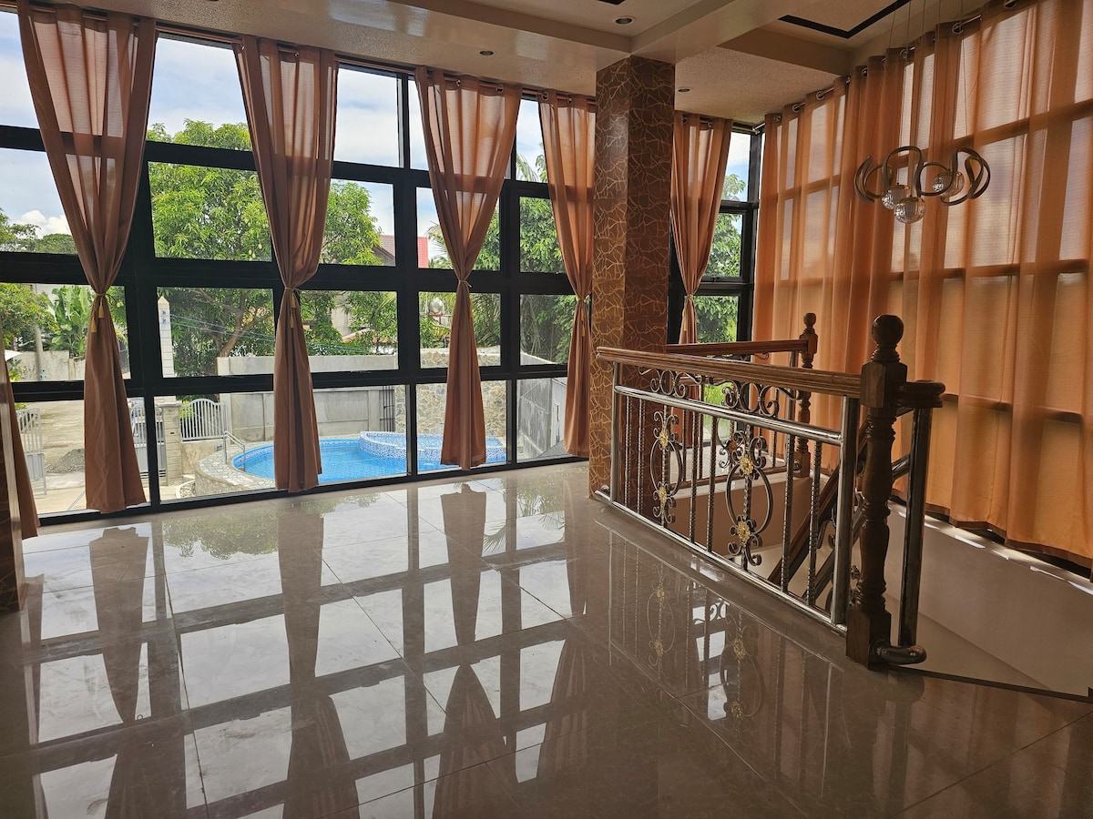 A bright and airy living space features large windows with flowing curtains, allowing natural light to illuminate the polished floors. A chandelier hangs from the ceiling, and a decorative railing borders the staircase, leading to an outdoor area with a pool visible through the glass.