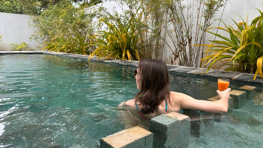 A tranquil swimming pool is surrounded by lush greenery and bamboo plants. Stone steps lead partially into the water, which reflects a calming blue hue. A guest enjoying a beverage is seen in the pool, contributing to a serene atmosphere.