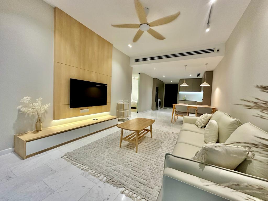 A modern living area is highlighted by a light-colored sofa positioned across from a wall-mounted television. A wooden coffee table sits atop a soft area rug. Ambient lighting is provided by ceiling fixtures, while a dining area with chairs is visible in the background.