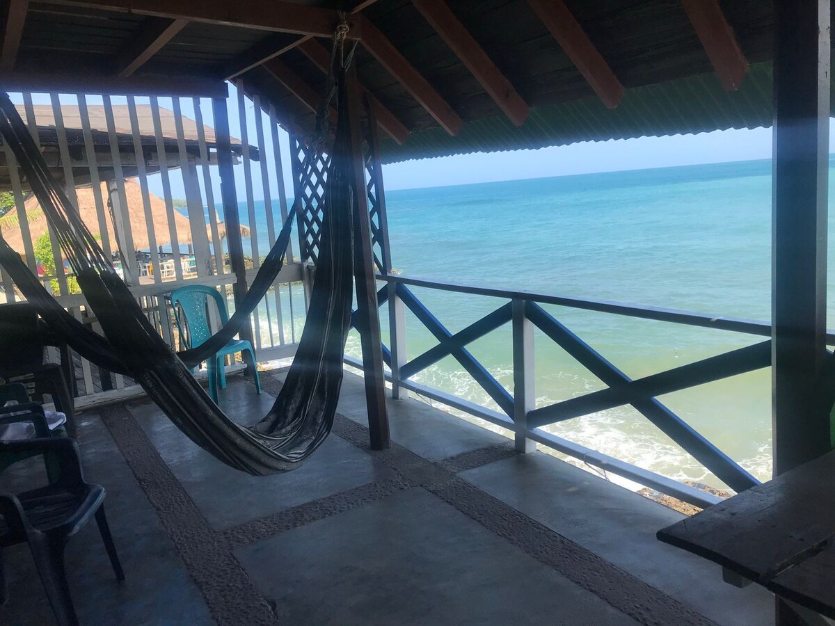 An open terrace showcases a sweeping view of the ocean, with waves gently lapping at the shore. A hammock hangs between supports, and plastic chairs are arranged nearby. Natural light illuminates the space, while the sound of the sea can be imagined.