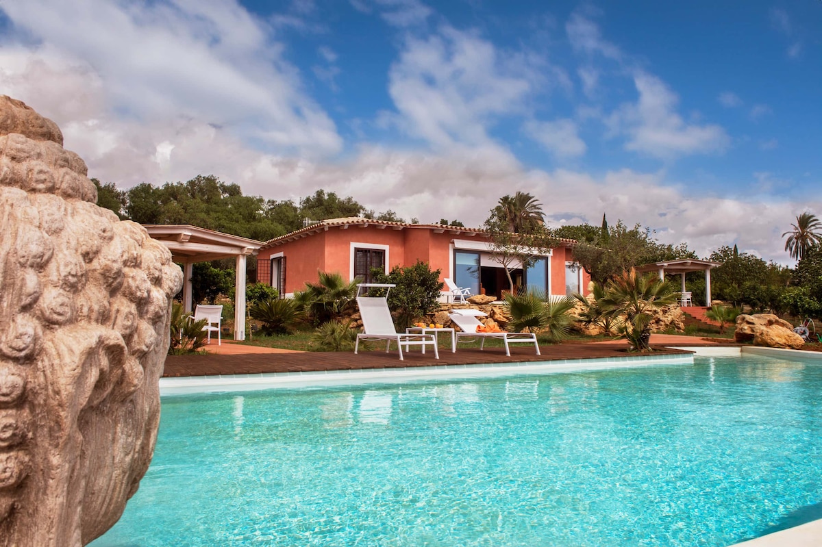 The villa is shown from a distance, framed by a clear blue pool in the foreground. Lush greenery surrounds the property, with white loungers positioned near the water. The structure features a pink exterior and large doors that open to the inviting outdoor space.
