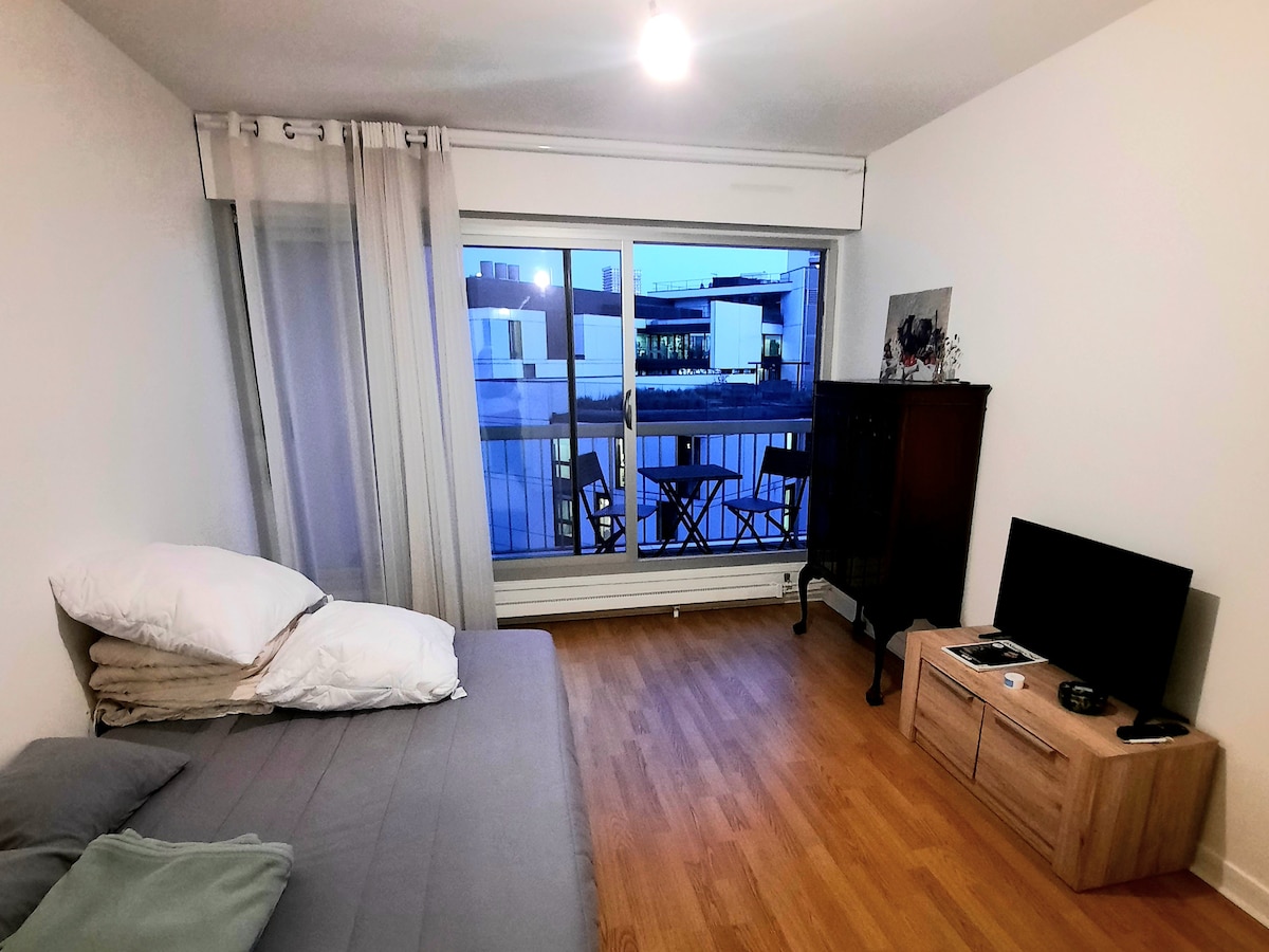 A spacious bedroom features a comfortable bed with layered bedding. A wooden TV stand supports a flat-screen TV. Light from a nearby balcony illuminates the room, where a small table and chairs are visible outside, creating an inviting outdoor space.