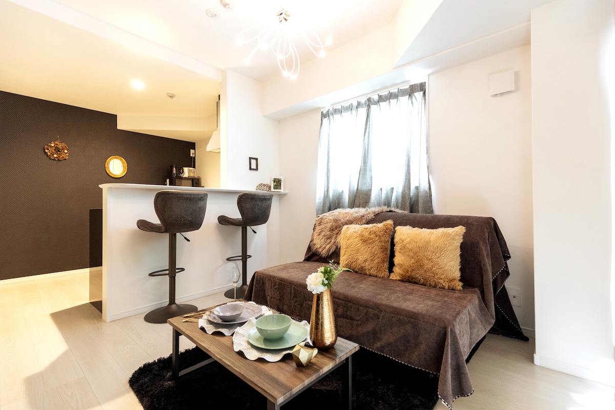 A modern living area features a stylish sofa adorned with decorative cushions. A sleek coffee table is set with dishware. The space is illuminated by a unique light fixture, with soft curtains framing the window. A kitchenette is visible in the background.