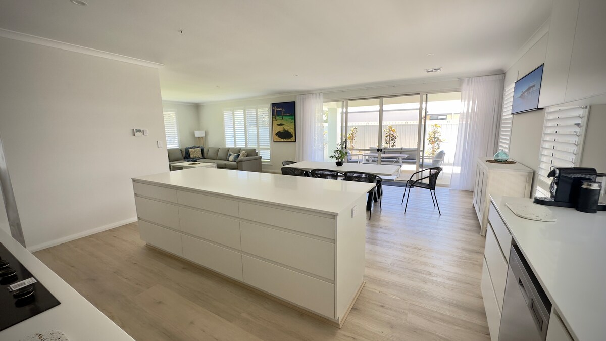 A spacious open-plan kitchen and dining area features a large white island with ample storage. Natural light fills the room through expansive glass doors, revealing a dining table and chairs, as well as a comfortable living area completed with modern furnishings.