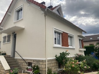 The exterior of a two-story house is visible, featuring a light-colored facade with decorative brick accents. Windows are bordered by white trim, and a set of stairs leads to the front door. Flower beds with various blooms are situated along the ground level.