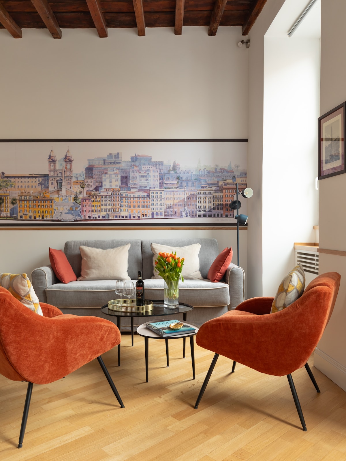 The living room showcases a gray sofa adorned with decorative pillows, flanked by two orange armchairs. A circular glass coffee table sits in the center, topped with a vase of flowers and a wine bottle. A large mural depicting a cityscape is displayed on the wall behind.