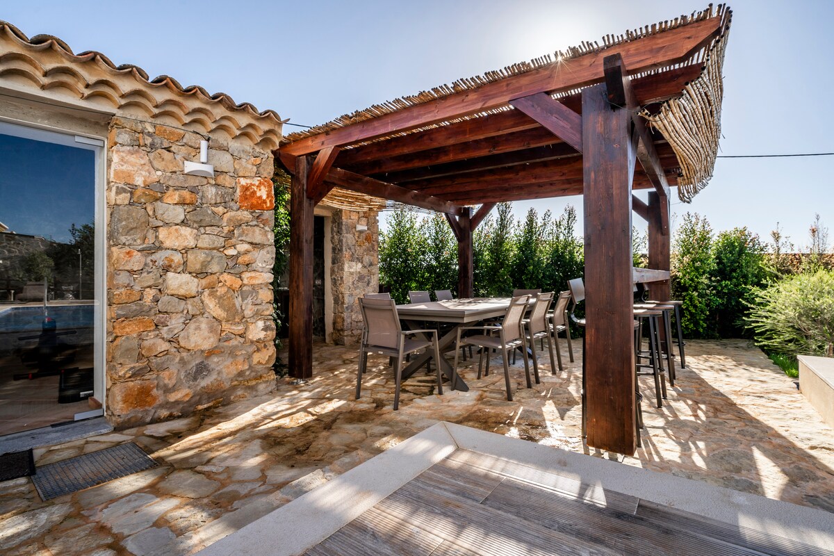 A shaded outdoor dining area is featured under a wooden pergola, supported by sturdy columns. A long rectangular table is surrounded by several chairs, inviting gatherings. Natural stone flooring leads the way, complemented by lush greenery in the background.