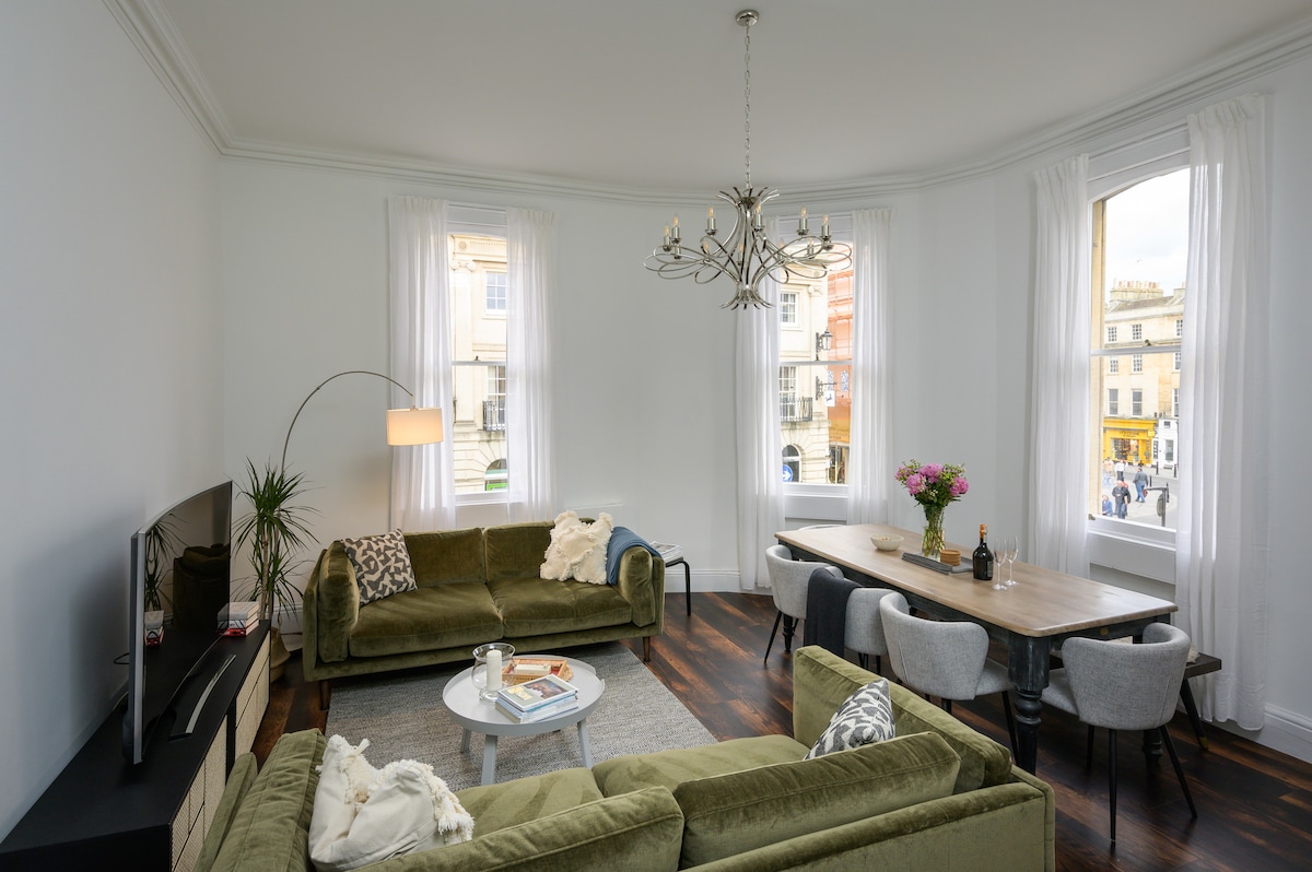 A bright living area is furnished with a pair of green sofas and a modern coffee table. A dining table with grey chairs is positioned nearby, and large windows with light curtains allow natural light to fill the space. A chandelier adds elegance to the room.