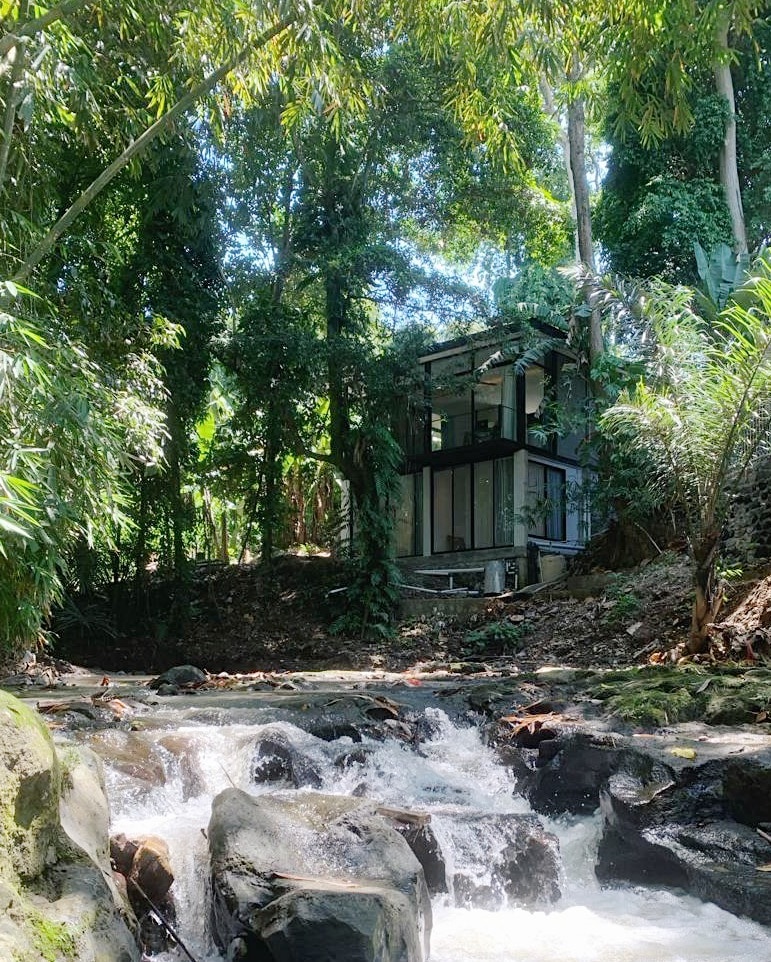 A modern two-story home is nestled among lush greenery, facing a gentle river. Tall trees and vibrant foliage surround the property, while a cascading water feature flows over smooth rocks in the foreground, enhancing the natural setting.