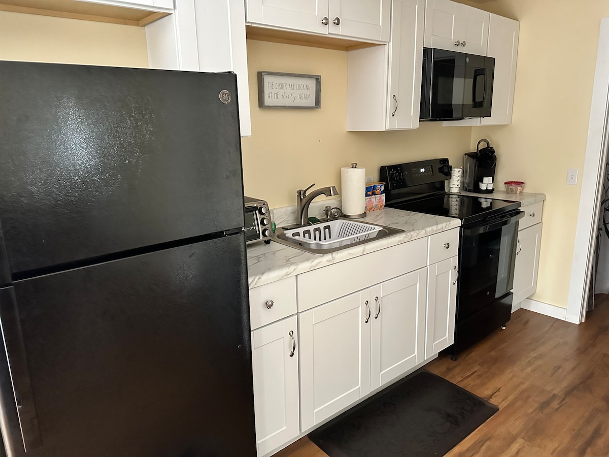 A modern kitchen is highlighted by sleek black appliances, including a refrigerator and oven. White cabinetry and a marble countertop provide ample workspace. A sink sits beneath a window, and essential kitchen items are neatly arranged on the countertop.