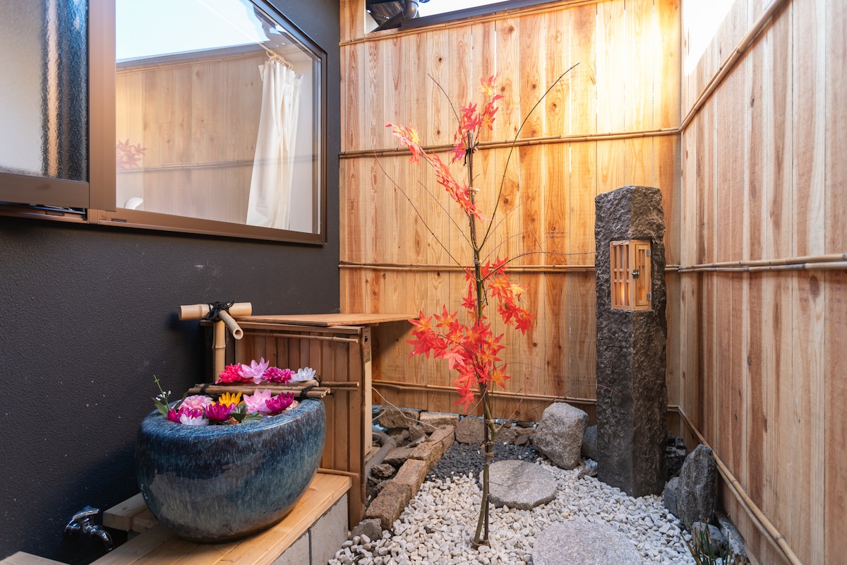 A serene nook features a stone water feature beside a round basin filled with colorful flowers. Smooth pebbles cover the ground, and a single maple tree with striking red leaves stands against a backdrop of wooden walls and a softly illuminated window.
