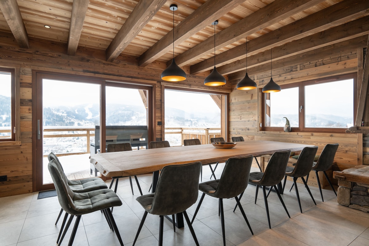 A spacious dining area is highlighted by a long wooden table surrounded by dark green chairs. Large windows allow natural light and mountain views, while industrial-style pendant lights hang above the table, creating a warm environment for meals.