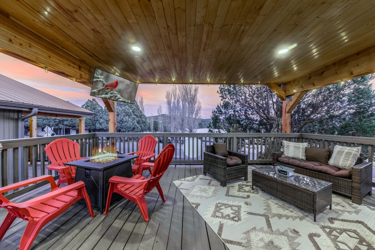 A spacious, covered deck features red Adirondack chairs surrounding a fire pit. A sectional sofa and coffee table create a cozy seating area. Striking mountain views are visible beyond the railing, with soft light illuminating the scene, enhancing the inviting outdoor space.