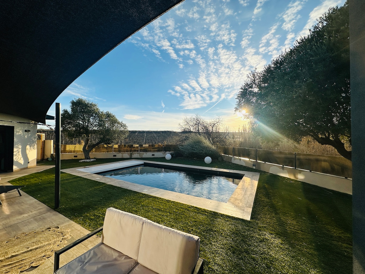 A tranquil outdoor space features a shaded area with lounge seating overlooking a heated pool. The pool is surrounded by manicured grass and visually connected to a serene landscape, with scattered trees and a clear sky reflecting the evening light.