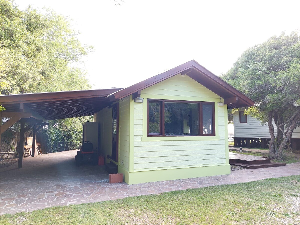 A charming yellow exterior of the house is showcased, with a gabled roof and large windows. An outdoor patio area is visible, featuring stone flooring and a grassy yard, surrounded by trees that provide greenery and shade.
