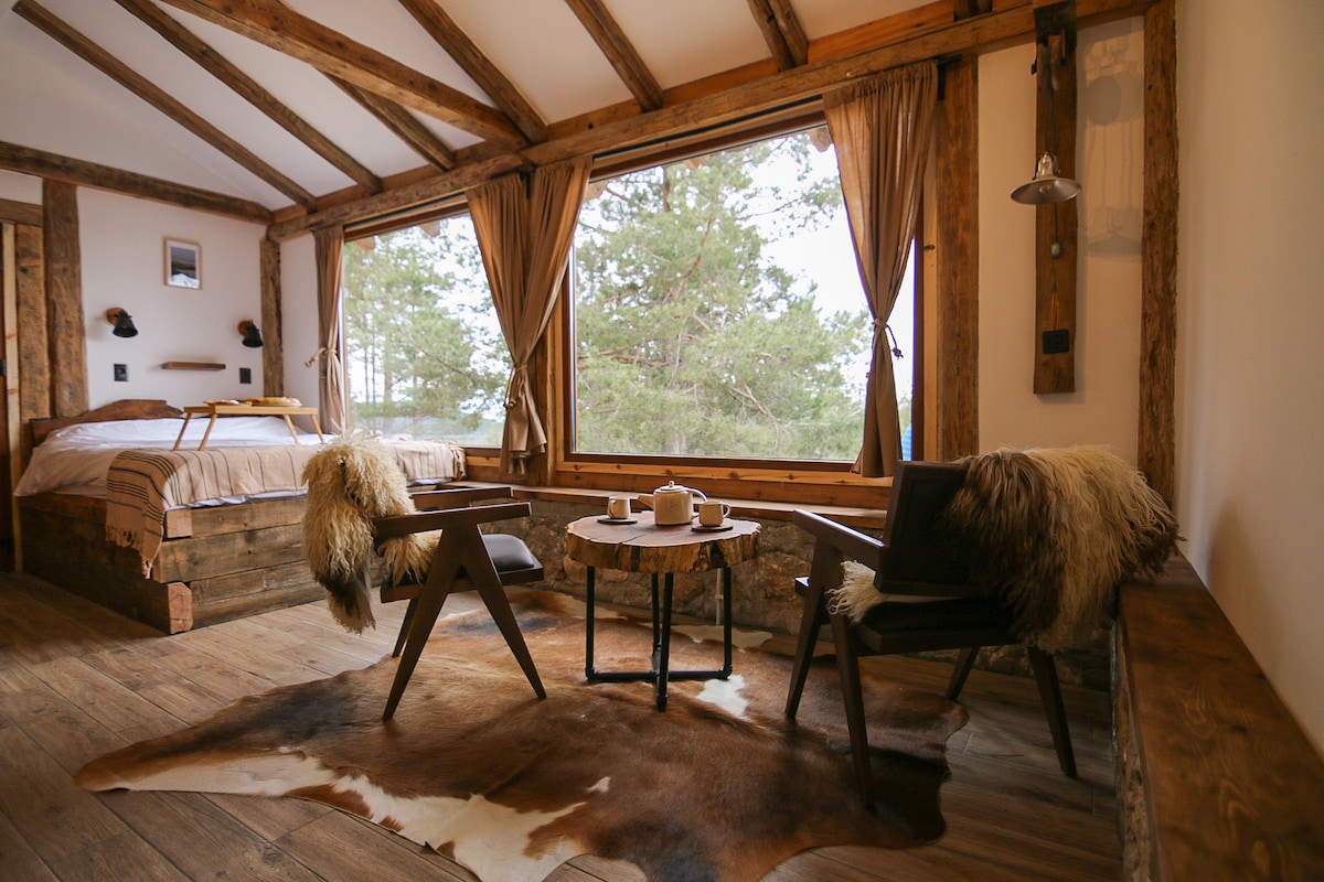 A cozy sleeping area features a spacious bed framed by wooden beams. Two chairs with soft throws are positioned around a small table, providing a comfortable seating arrangement. Large windows offer views of the surrounding nature, with soft curtains framing the outdoor scenery.