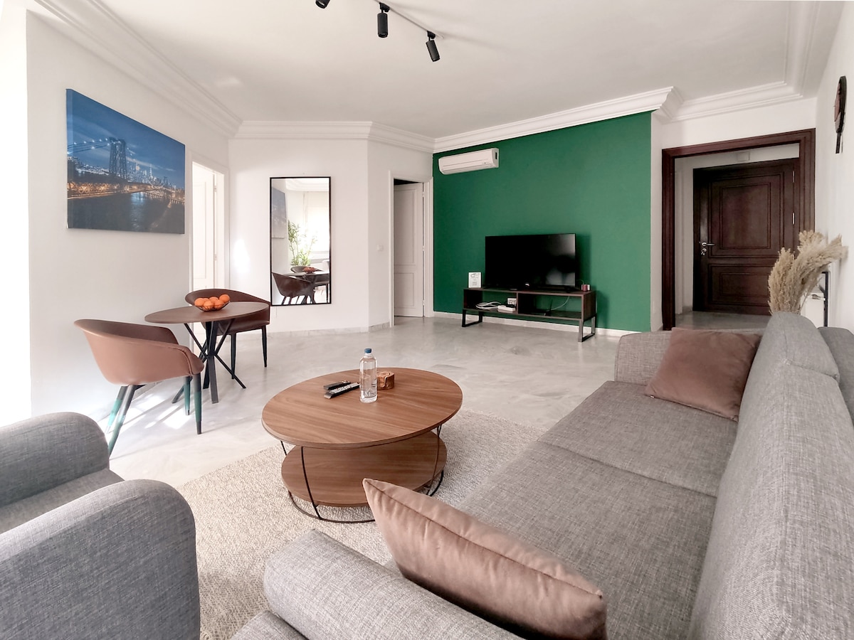 A modern living area features a comfortable gray sofa with decorative pillows, a round wooden coffee table, and a dining table set for four. A green accent wall adds depth, while a flat-screen TV is positioned on a light-colored media console. Large windows allow natural light to enter.