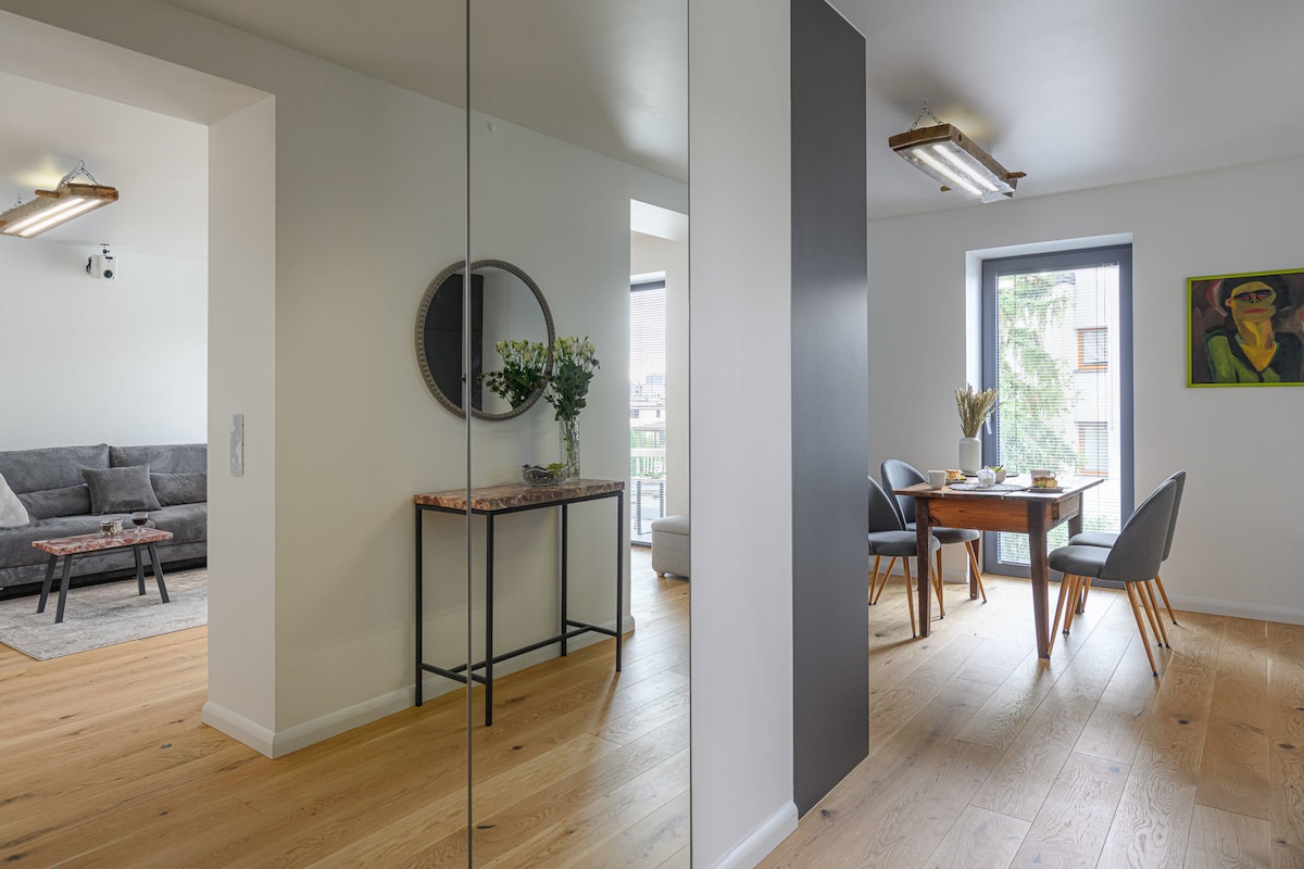 An open-plan living area is seen, featuring a cozy lounge space with a gray sofa and a dining table for four. Natural light fills the room through large windows, highlighting the wooden flooring and modern decor. Fresh flowers are placed on the table, adding a touch of warmth.