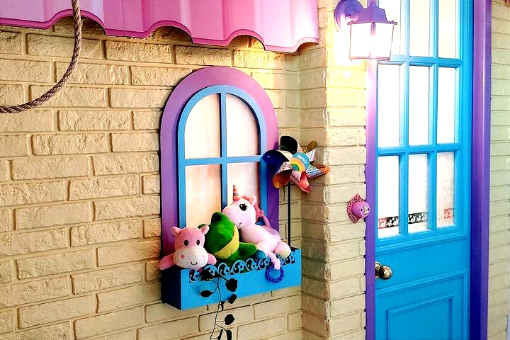 A colorful window box features plush toys arranged playfully, with a bright blue window framed by a charming pink awning. The textured brick wall adds a whimsical touch to the cheerful entrance, inviting all to explore the fun-filled interior.