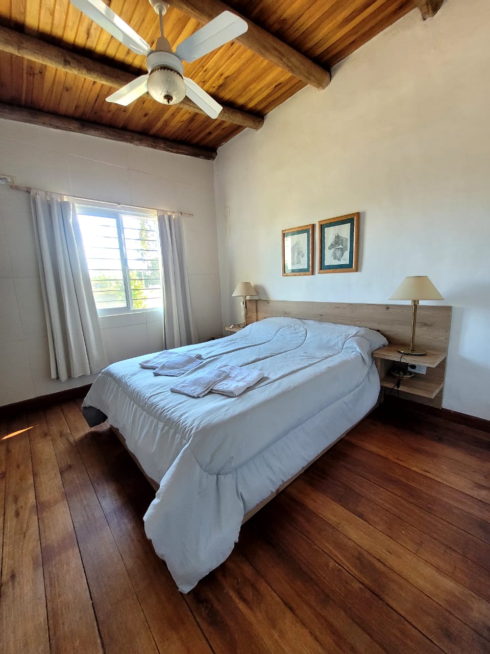 A well-lit bedroom features a spacious bed with neatly arranged linens. Two bedside lamps are positioned on either side of the bed. The wooden ceiling and floor add warmth to the space, while a window allows natural light to brighten the room.