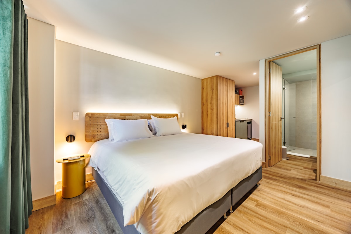 A spacious bedroom is highlighted by a large bed with crisp white bedding. Wooden paneling adds warmth, while a green curtain frames a window. A stylish side table is next to the bed. An open doorway leads to a modern bathroom area.