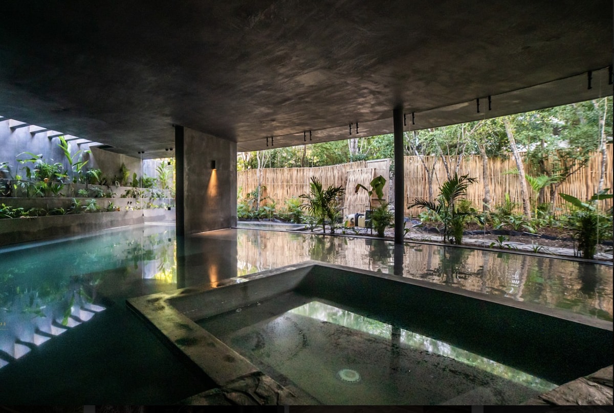 A serene pool area is surrounded by tropical greenery, with large windows offering an open view to the outdoors. The sleek water surface reflects the surrounding foliage, while a hot tub is integrated into the design. Soft lighting creates a calm atmosphere, enhancing the sense of tranquility.