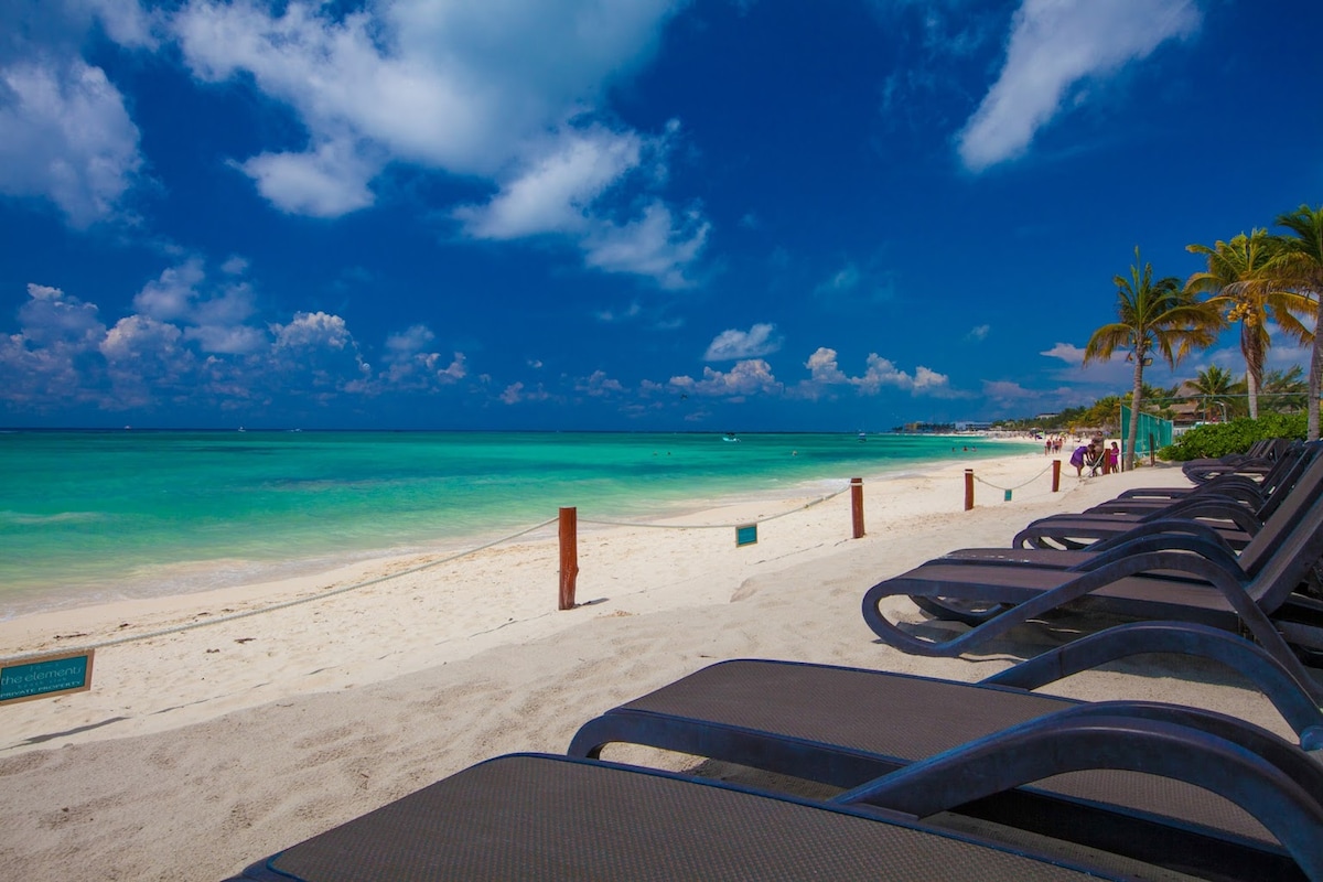 A stretch of sandy beach is shown, lined with lounge chairs facing the turquoise ocean. Palm trees are visible in the background, along with a clear blue sky dotted with clouds. The gentle waves create a serene atmosphere suitable for relaxation.