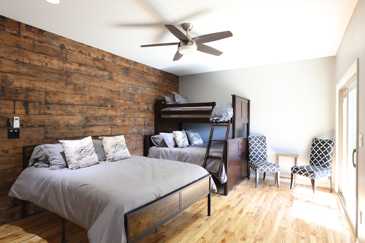 A spacious bedroom features a rustic wood accent wall and a ceiling fan. A bunk bed is placed against the wall, complemented by a large bed with soft gray bedding. Two patterned chairs are positioned near a set of sliding glass doors, allowing natural light to enter.
