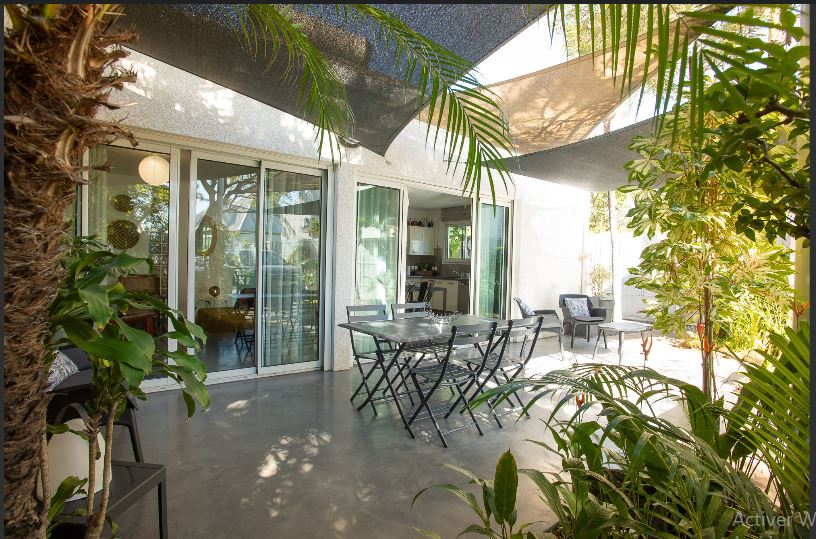 An outdoor terrace is surrounded by lush greenery, featuring a dining table with several chairs. Shade sails provide protection from the sun, while sliding glass doors open to an interior space visible in the background.