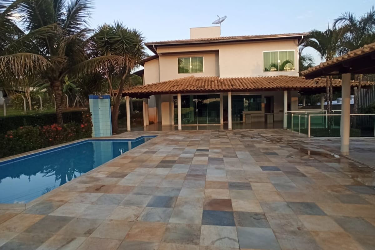 A two-story house is visible in the background, featuring a roof with terracotta tiles. In the foreground, a tiled patio surrounds a private swimming pool, with palm trees providing greenery. A covered outdoor area with a wooden roof is also present, offering shaded leisure space.