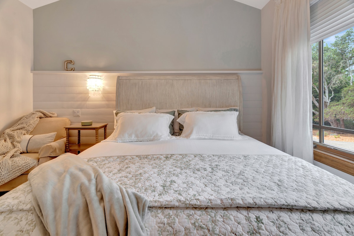 A spacious bedroom with a comfortable king bed adorned in soft linens. A cozy reading chair sits nearby, complemented by elegant side tables. Natural light floods in through the large window, showcasing views of the surrounding trees.