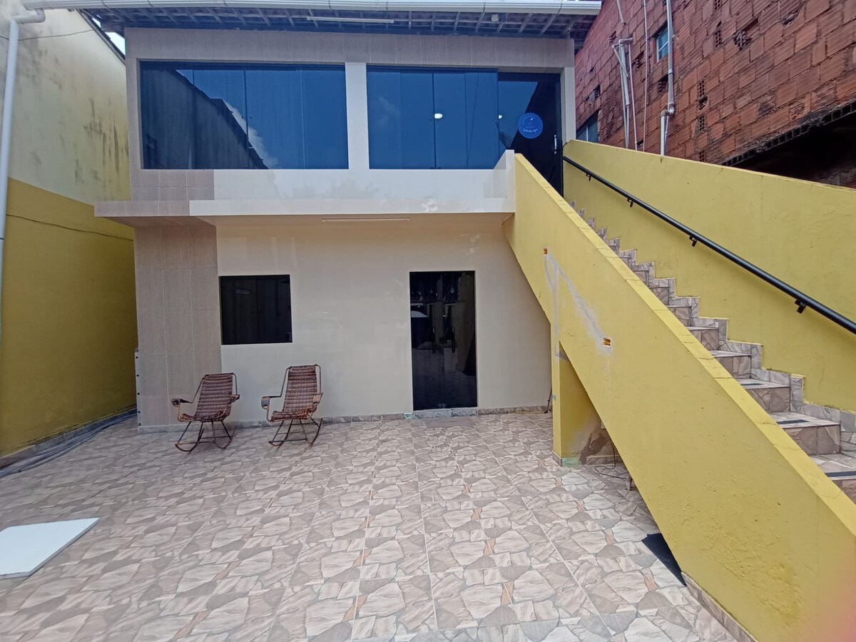 An outdoor area is presented with a tiled floor and a staircase leading to an upper level. Two chairs are positioned on the ground, and large windows provide natural light to the interior behind. A bright yellow wall adds a distinctive touch to the space.