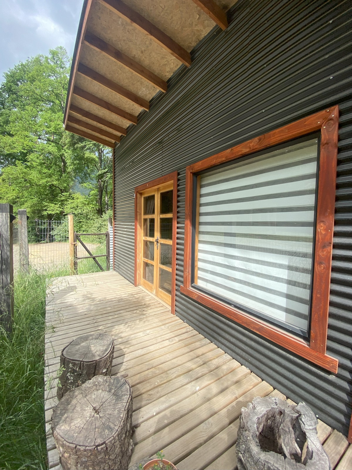 An outdoor wooden deck features two rustic logs as seating, positioned in front of a large window with contrasting light and dark striped curtains. The textured exterior walls display varying shades of dark material, complemented by wooden trim, surrounded by lush greenery.