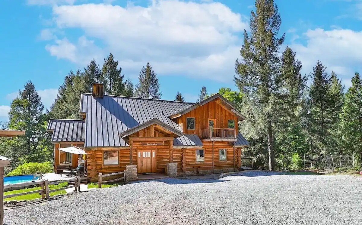 A custom-built log cabin is set amidst a natural landscape. The structure features a combination of wood and metal roof with large windows. Surrounding greenery includes tall trees, and a gravel driveway leads to the entrance, creating a serene and spacious outdoor environment.