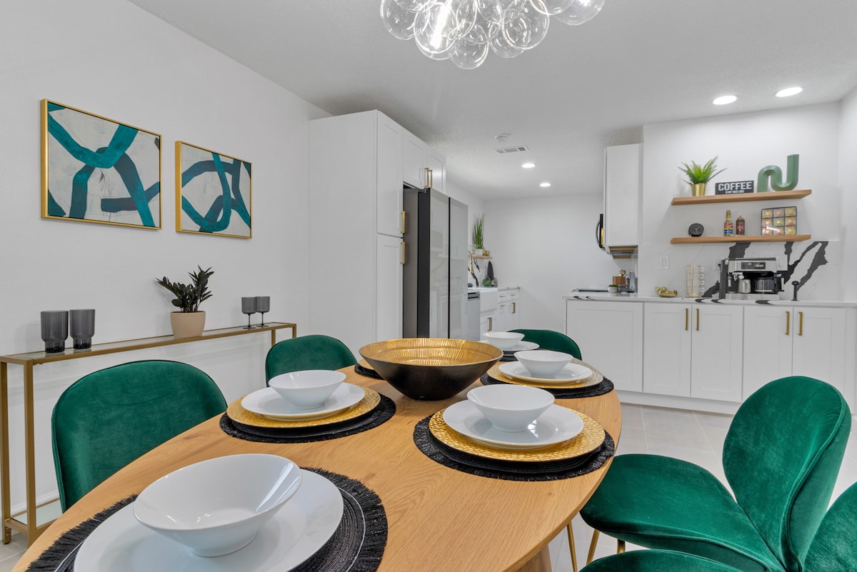 A modern dining area features a round wooden table set for six, accented with green chairs and black placemats. Two abstract art pieces adorn the white walls, accompanied by stylish lighting overhead. A sleek kitchen cabinetry can be seen in the background, enhancing the home's contemporary look.