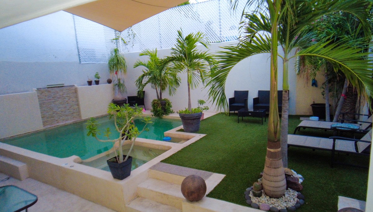 A tranquil outdoor space features a shimmering pool surrounded by lush greenery and potted plants. Comfortable lounge chairs are positioned on a grassy area, providing relaxation spots, while a tasteful stone accent wall adds a natural element to the serene setting.