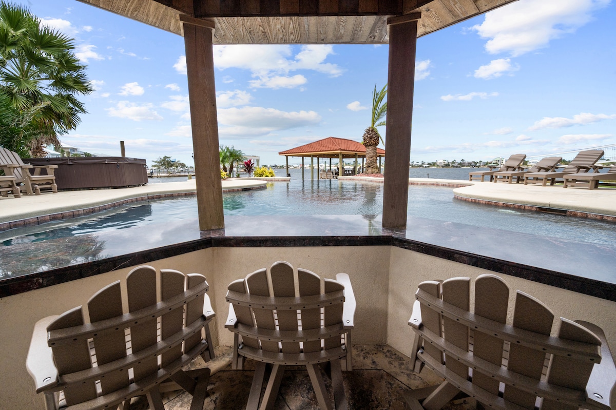 A shaded seating area features three adirondack chairs overlooking a private heated pool. The pool reflects the clear sky, with a wooden pavilion situated in the background. Lush palm trees and vibrant landscaping contribute to the serene atmosphere.